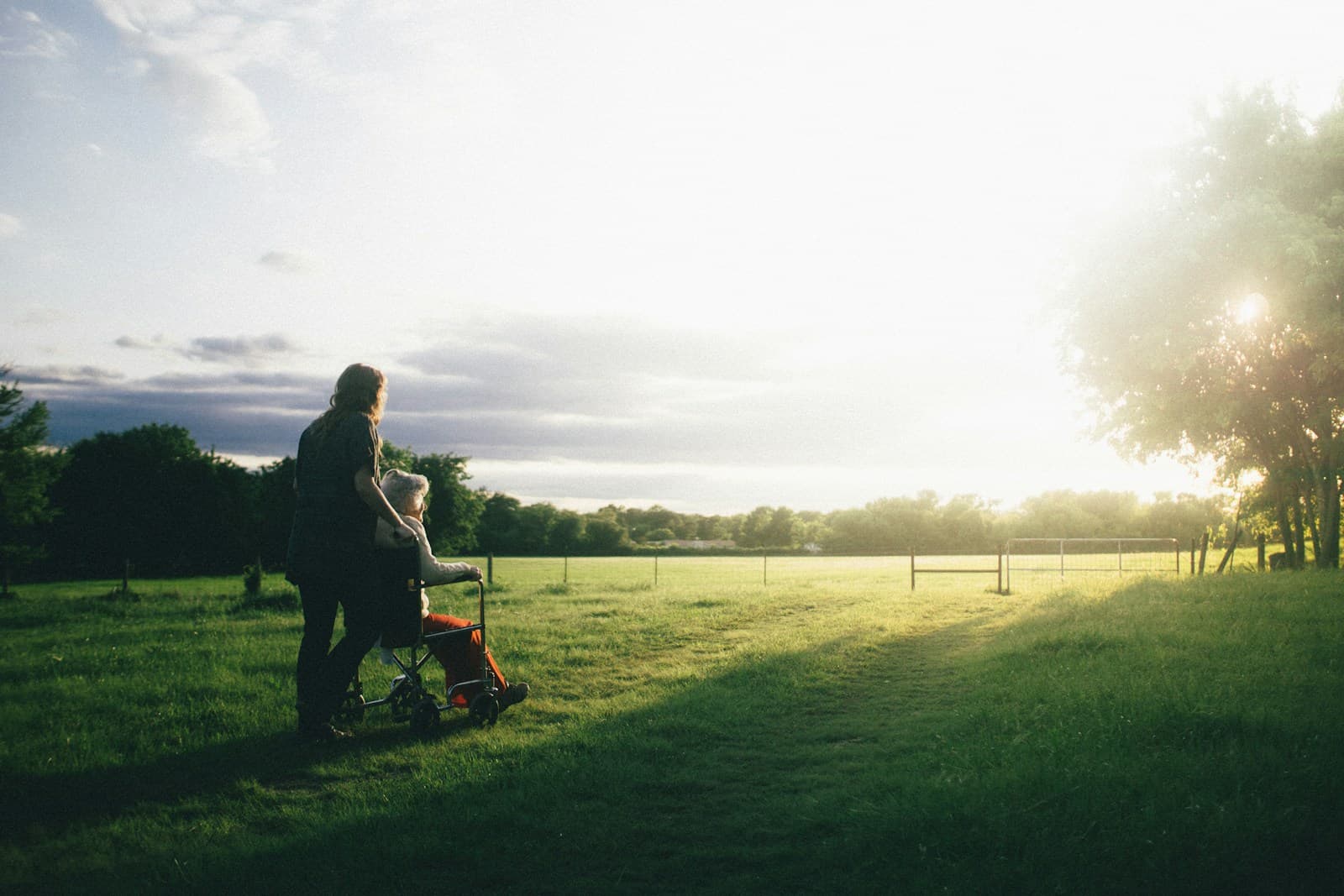 A carer pushing an elderly woman in a wheelchair across a sunlit meadow